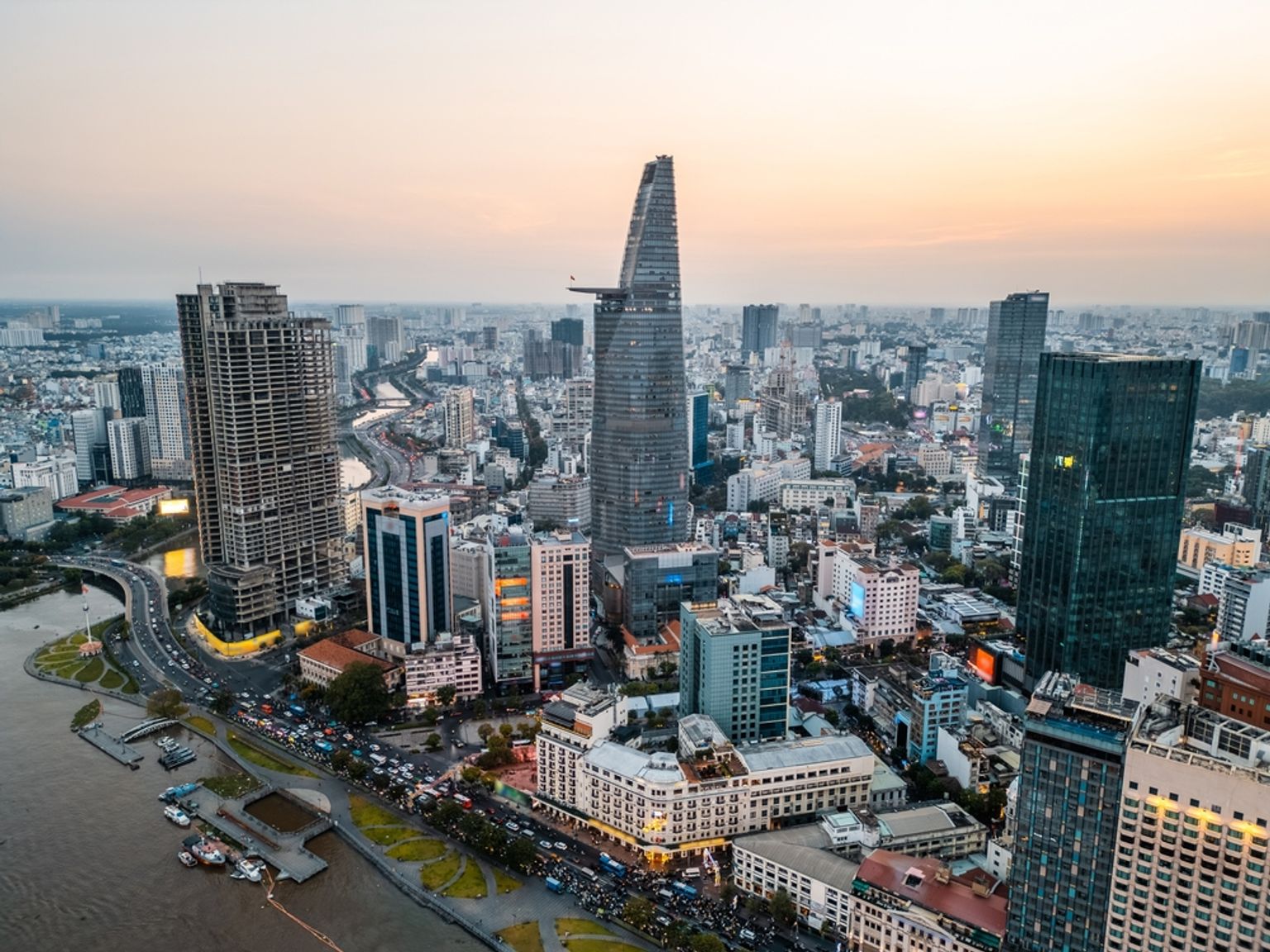 Luftfoto af Ho Chi Minh City ved skumring, med Bitexco Financial Tower, skyskrabere, en flod og travle gader.