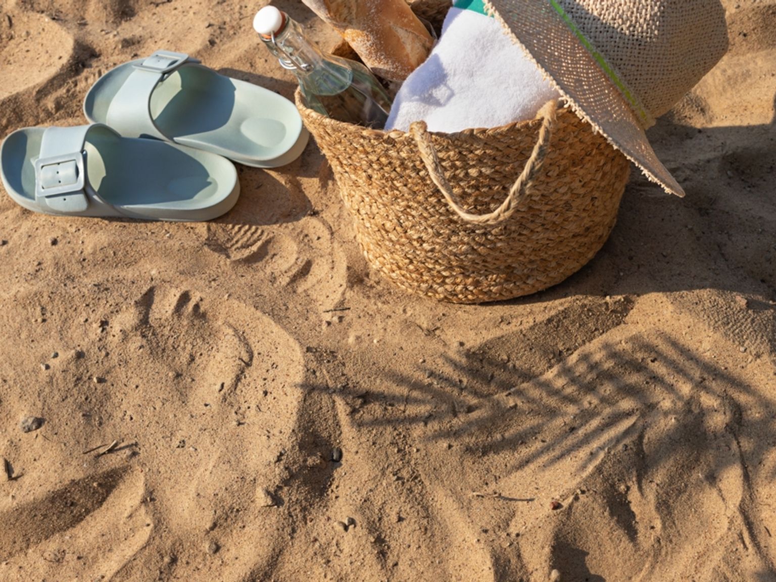 Ett par ljusblå sandaler och en strandväska av halm med en hatt och en handduk på sanden, med en palmsskugga.
