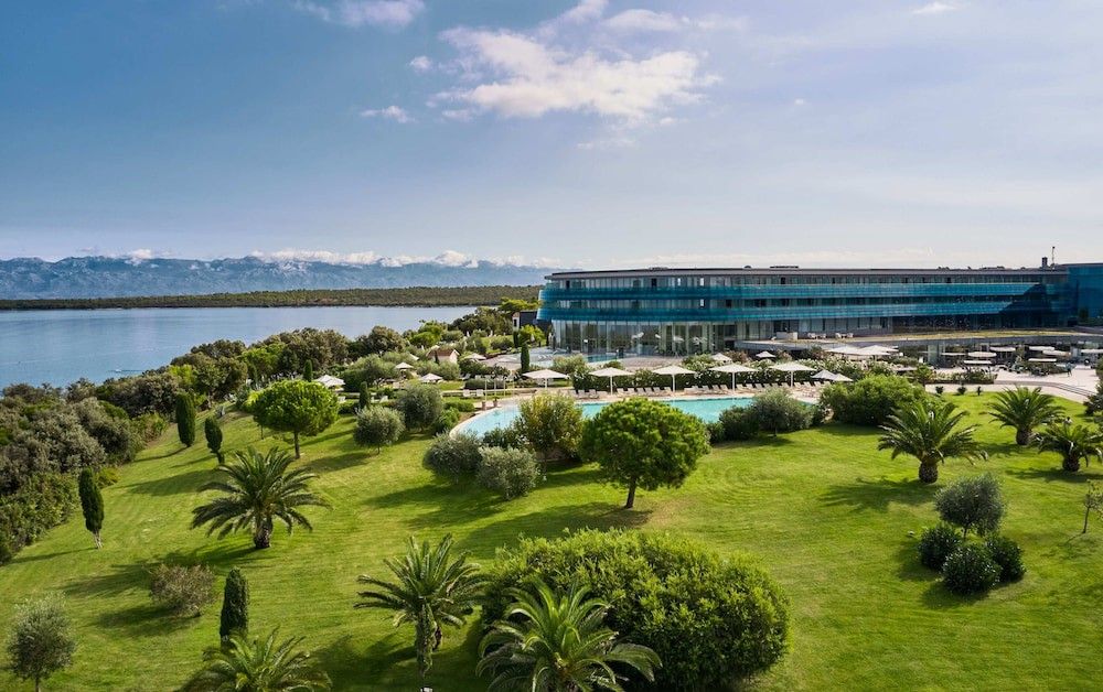 Luftfoto av et moderne resort-hotell med et basseng, grønne hager, en bukt og fjerne fjell