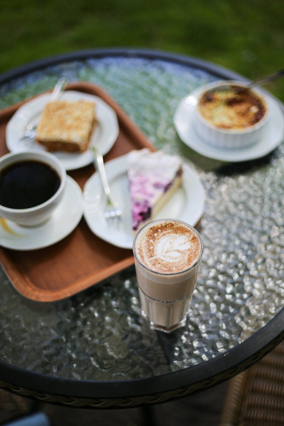 Et glas latte med blad-lattekunst, en kop sort kaffe, et stykke cheesecake, en firkantet kage og crème brûlée på et glasbord udendørs.