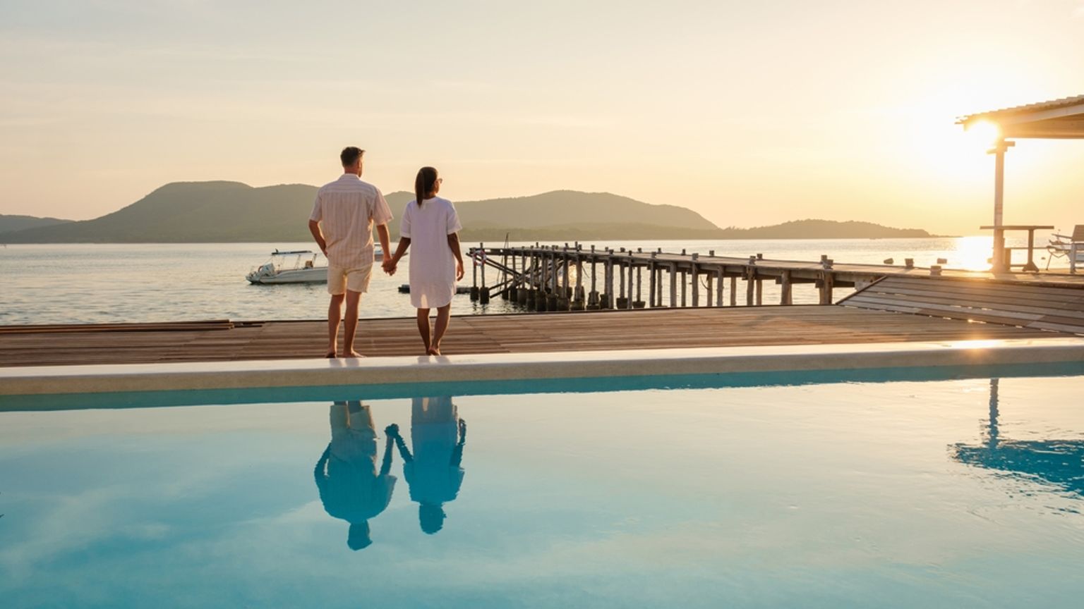 Pareja cogida de la mano junto a una piscina infinita, contemplando la puesta de sol sobre el mar y un muelle.