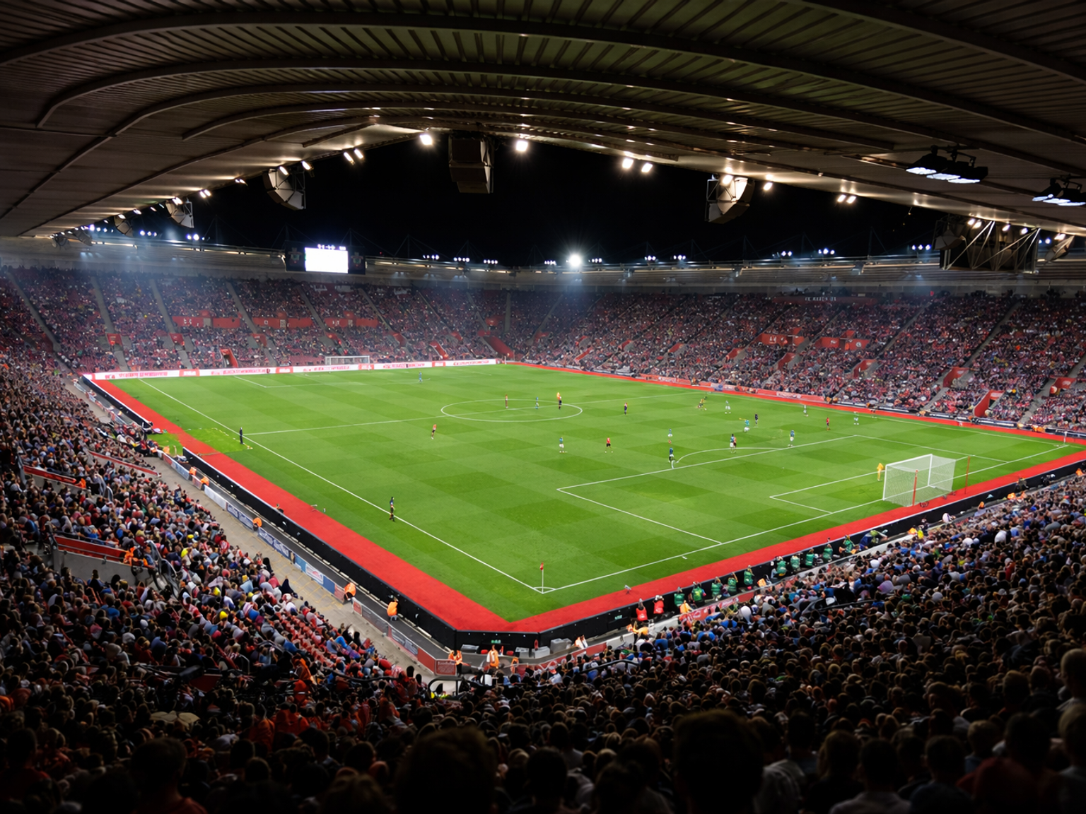 En natudsigt over et fodboldstadion fyldt med tilskuere, der ser en kamp på en stærkt oplyst grøn bane.