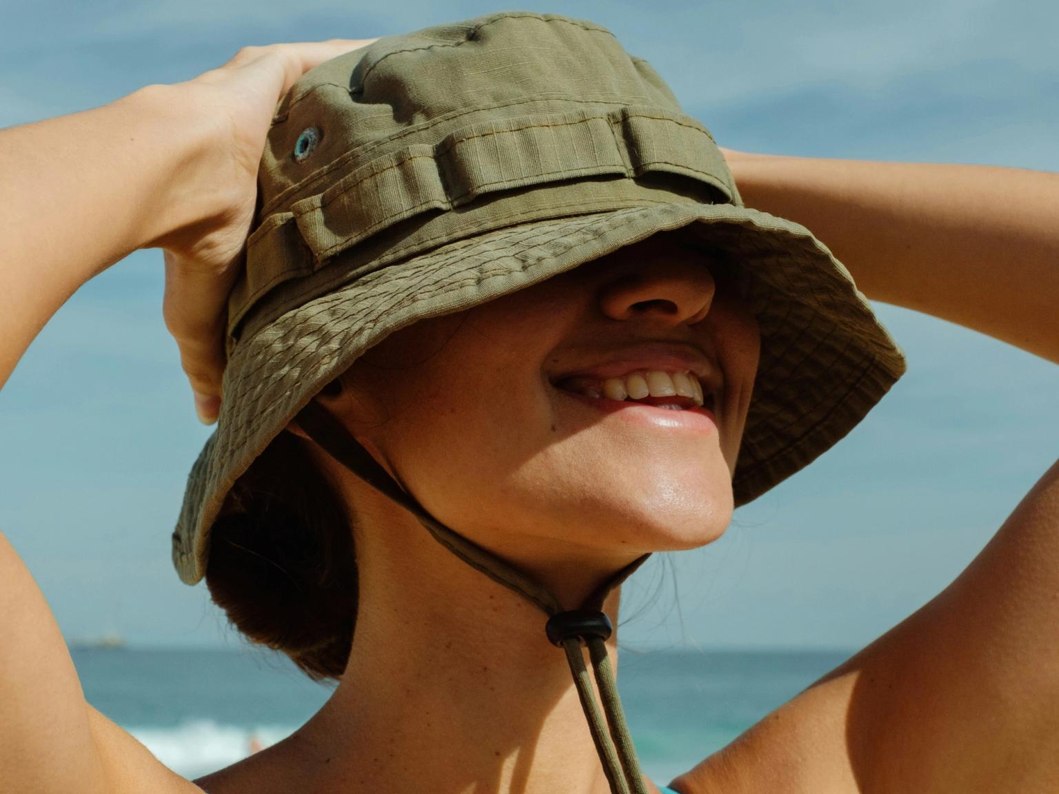 Lächelnde Person mit olivgrünem Fischerhut und blauem Badeanzug an einem sonnigen Strand.