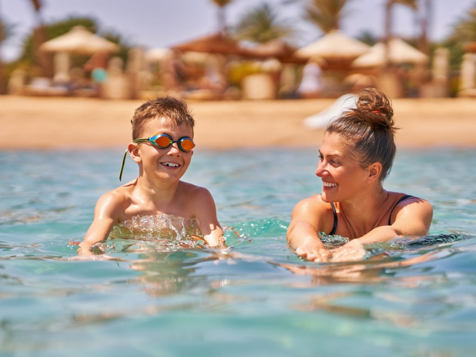 Ein lächelnder Junge mit Schwimmbrille und eine Frau schwimmen im Meer an einem Strandresort.