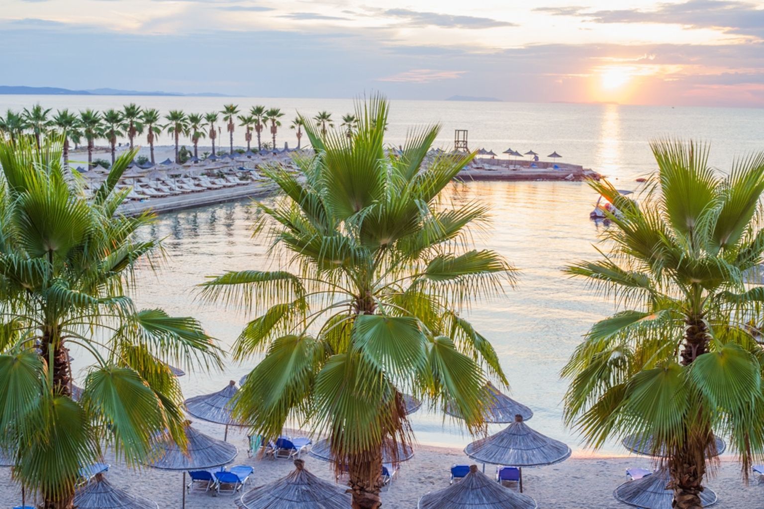 Solnedgang over en tropisk strand med palmer, stråparasoller og roligt hav.