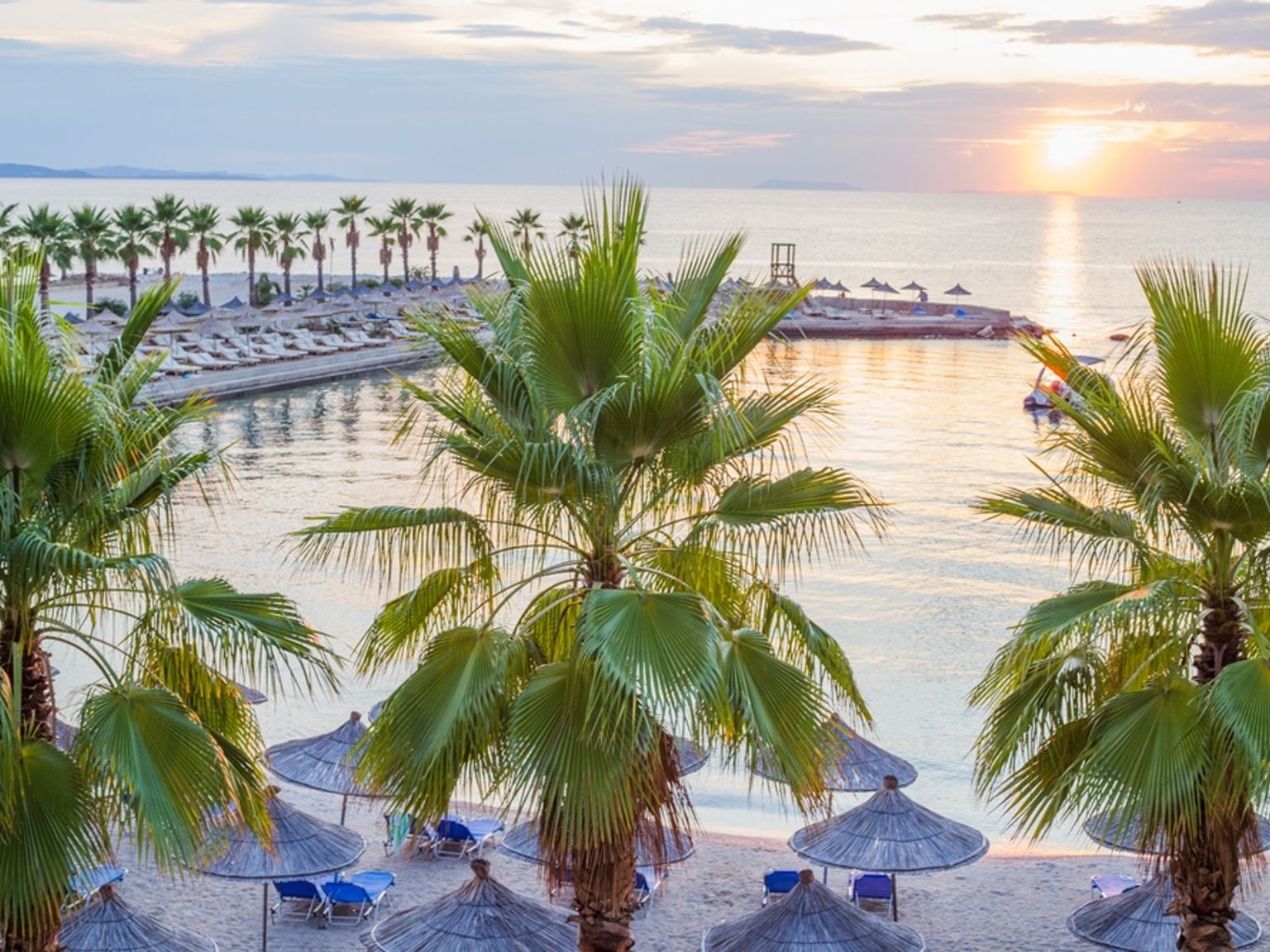 Solnedgang over en tropisk strand med palmer, stråparasoller og roligt hav.