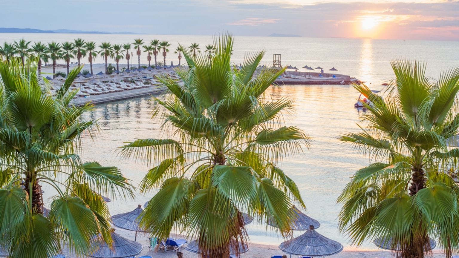 Solnedgang over en tropisk strand med palmer, stråparasoller og roligt hav.