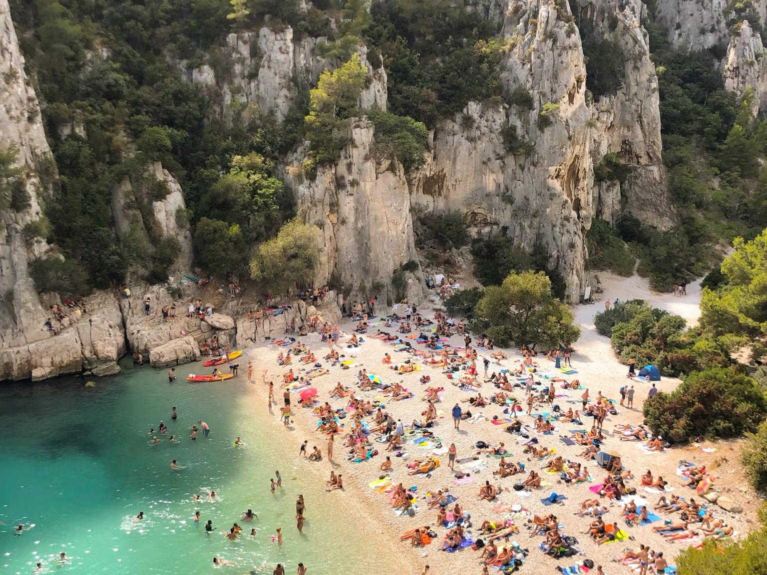 En fullsatt strand med människor som solar och badar, inbäddad mellan höga klippiga klippor och klart turkost vatten, Franska rivieran.