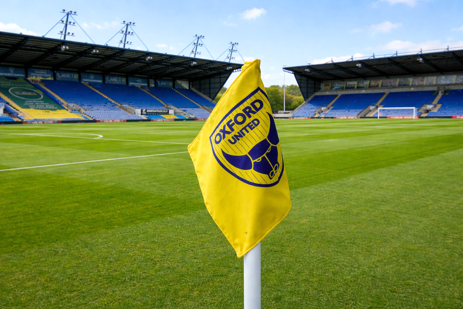 A yellow Oxford United corner flag on a green soccer field with empty blue stadium stands in the background.