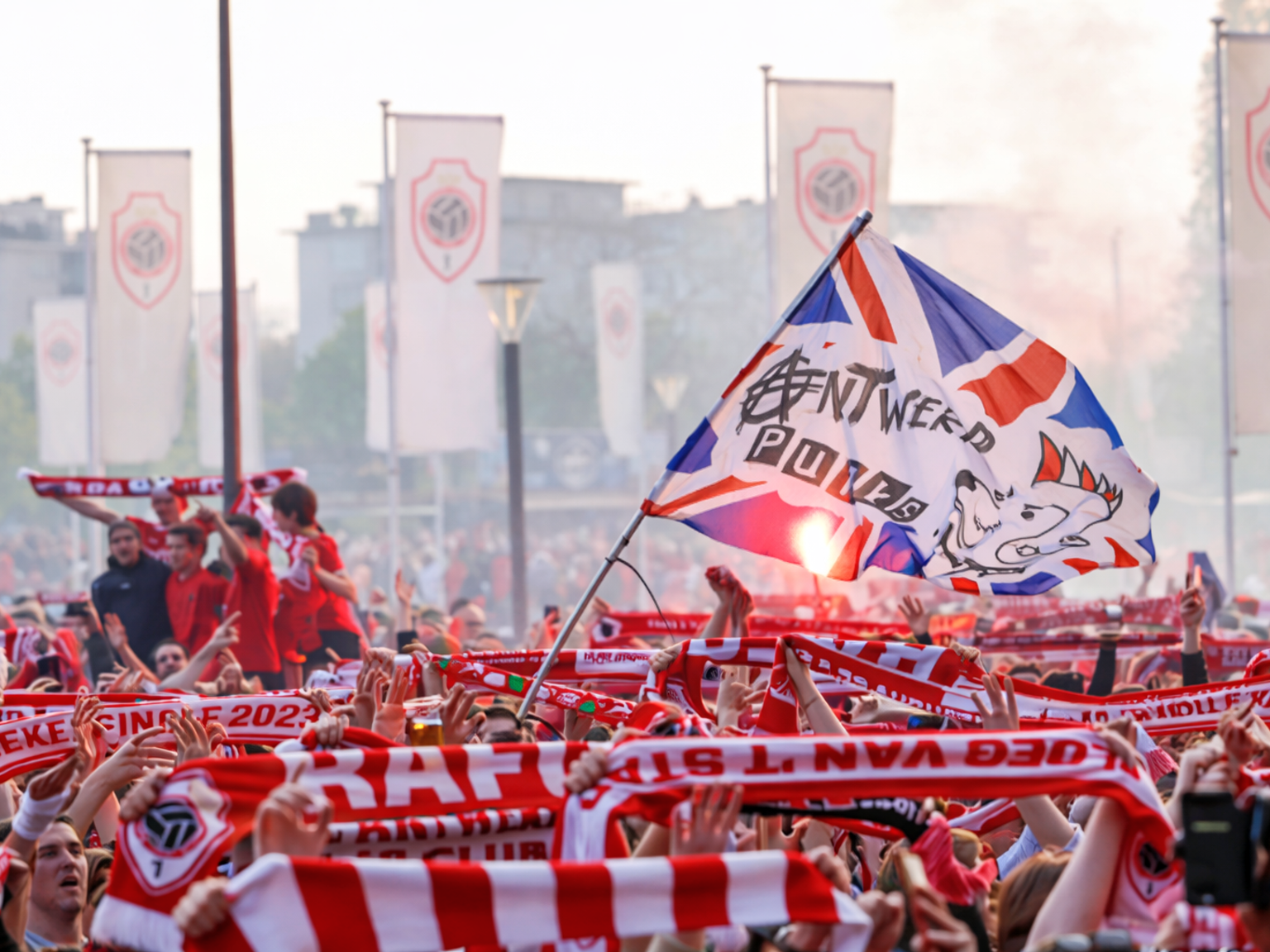 Eine große Menge von Fans des Royal Antwerp FC feiert begeistert, schwenkt rot-weiße Schals und eine Fahne mit der Aufschrift „Antwerp Power“, während roter Rauch im Hintergrund aufsteigt.