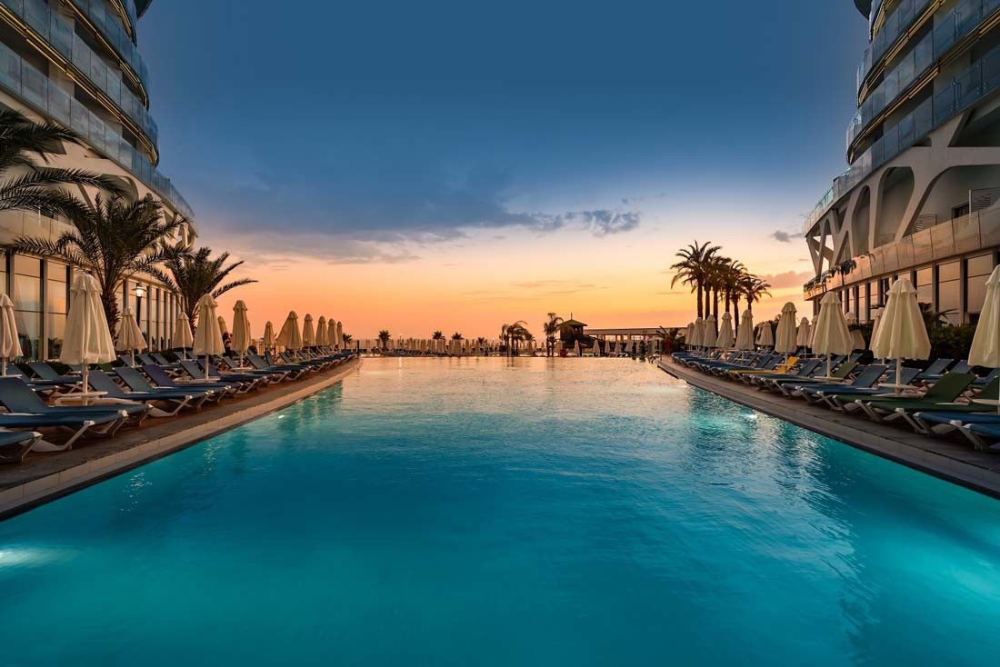 A long resort swimming pool at sunset, flanked by modern buildings, lounge chairs, and palm trees. Vikingen Infinity Resort and Spa, Alanya