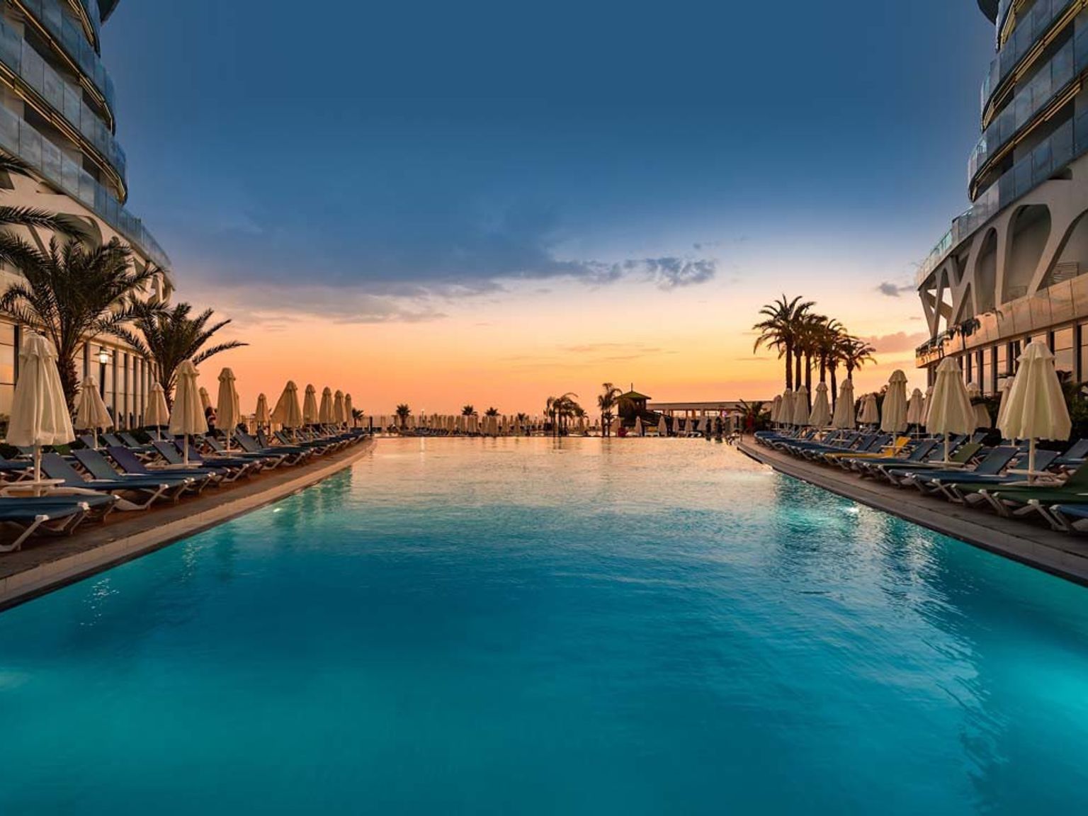 En lång resortsimbassäng vid solnedgången, kantad av moderna byggnader, solstolar och palmer. Vikingen Infinity Resort och Spa, Alanya