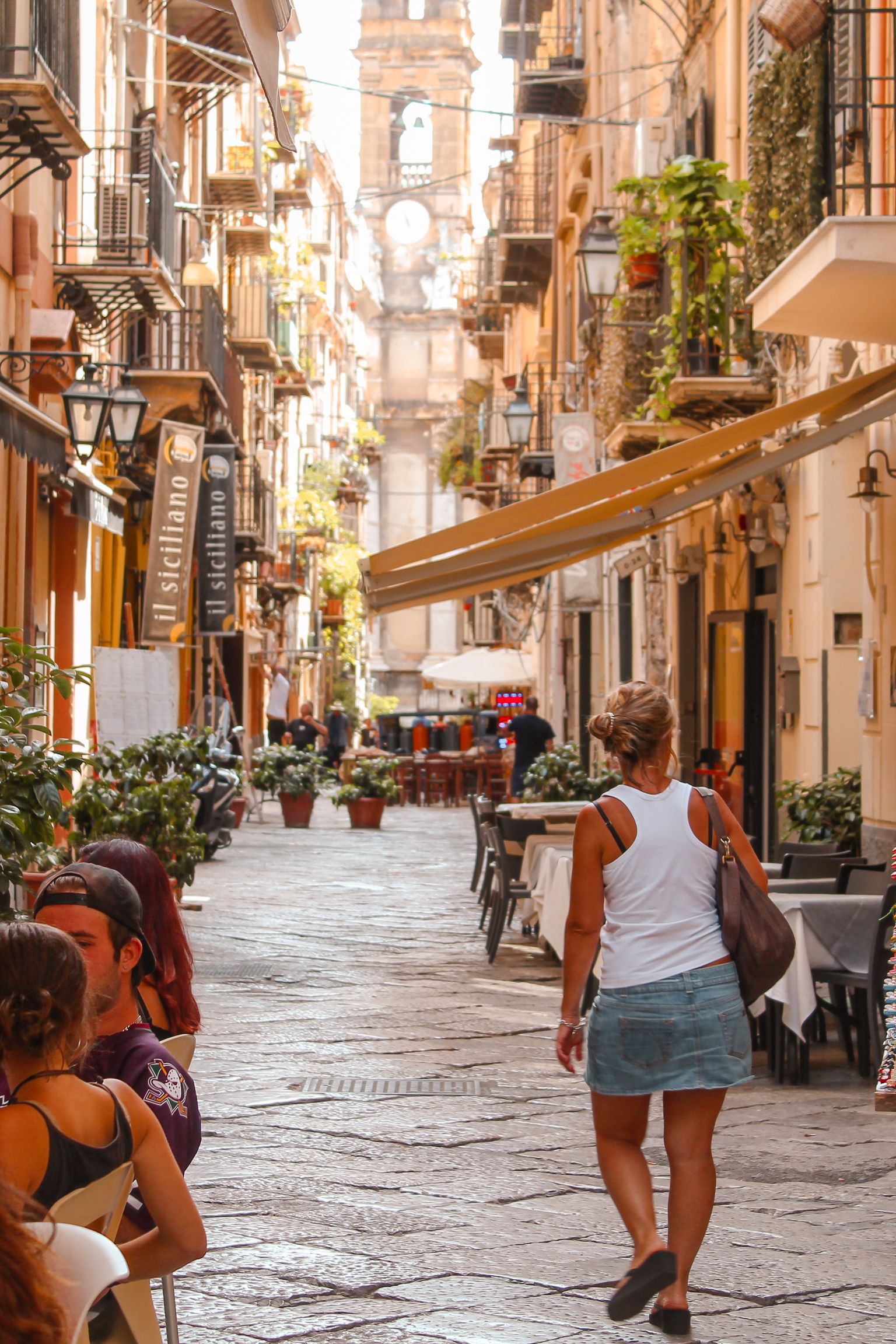 En livlig, smal gata med människor som äter på uteserveringar, en kvinna som går och en försäljare som säljer färgglada varor från en vagn, med ett avlägset torn.