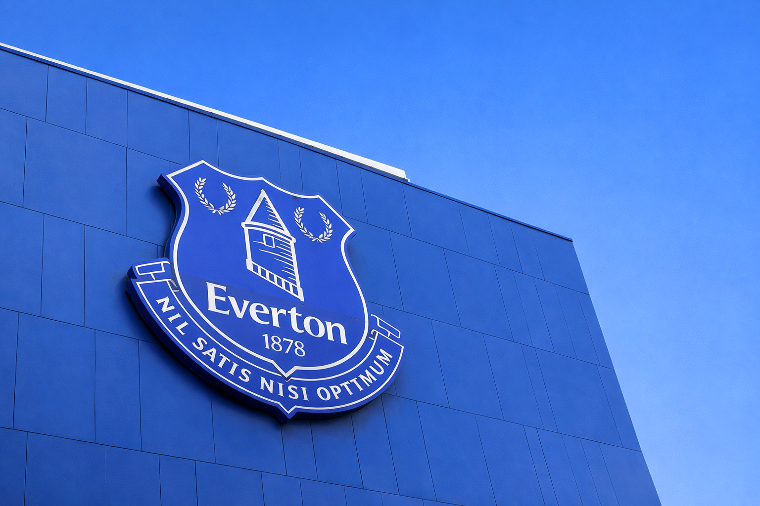 Uma grande bandeira do Everton FC acena sobre uma multidão de fãs de azul, muitos segurando cachecóis "EVERTON".