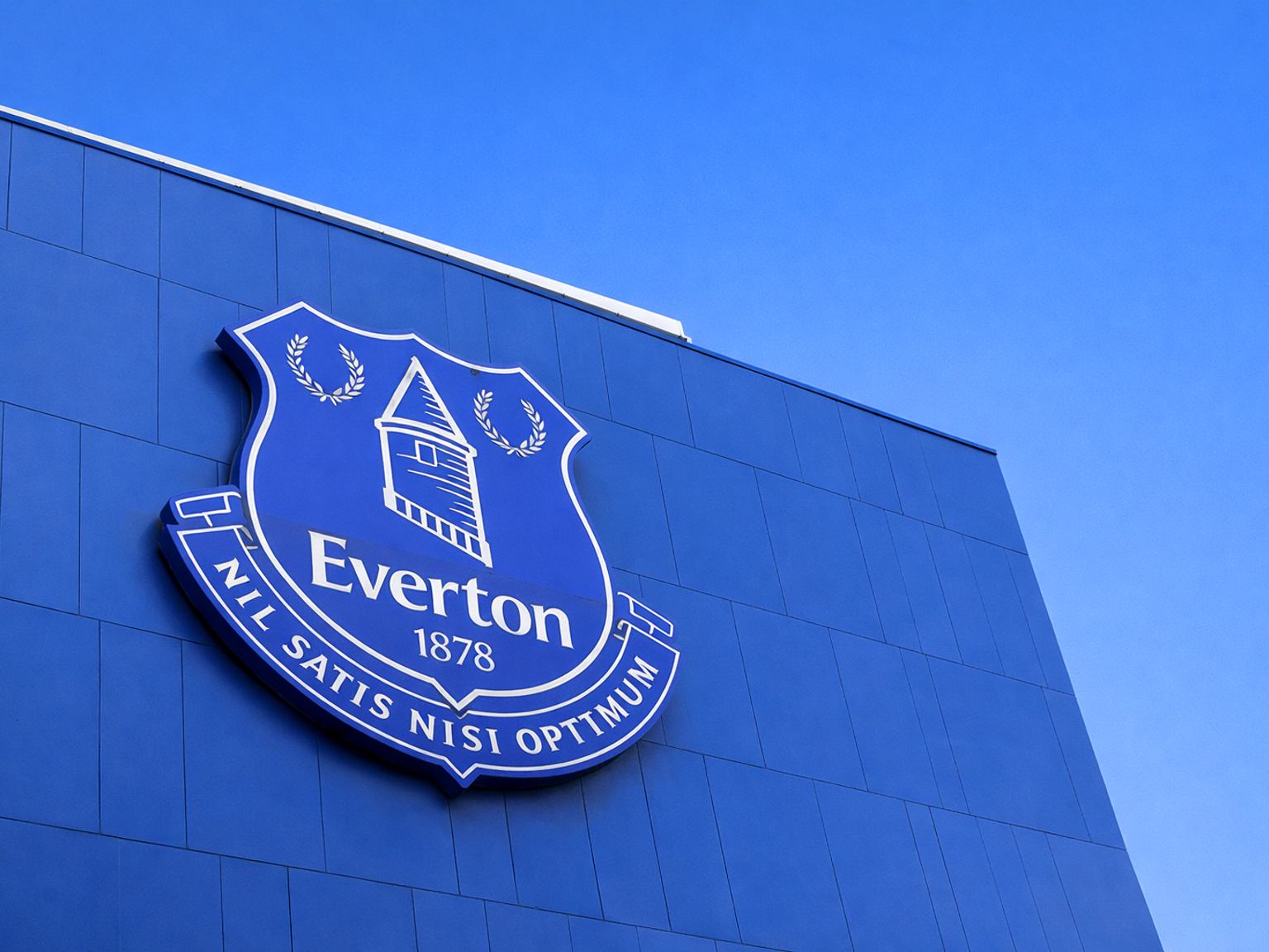 Uma grande bandeira do Everton FC acena sobre uma multidão de fãs de azul, muitos segurando cachecóis "EVERTON".