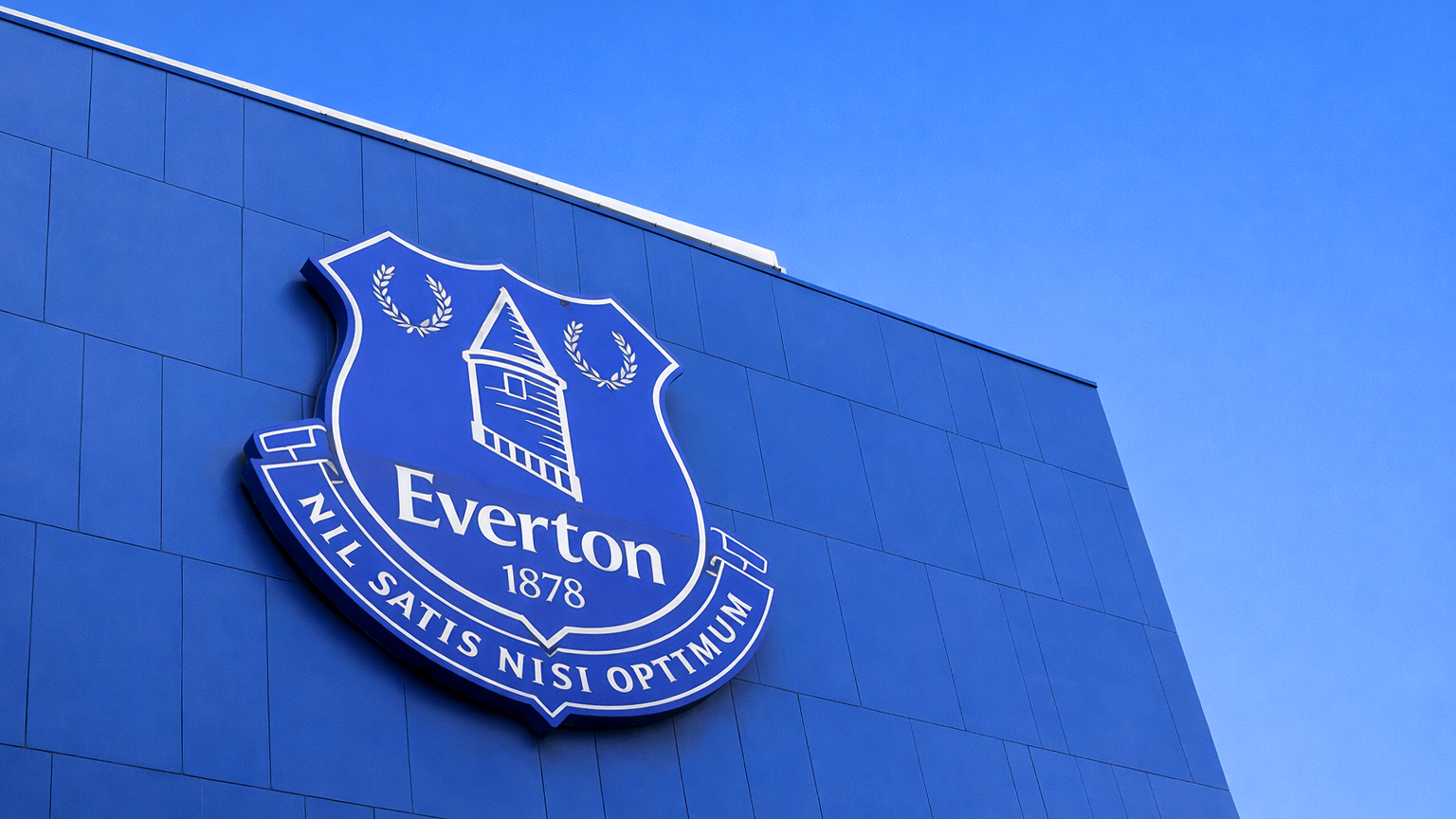 A large Everton FC flag waves over a crowd of fans in blue, many holding "EVERTON" scarves.