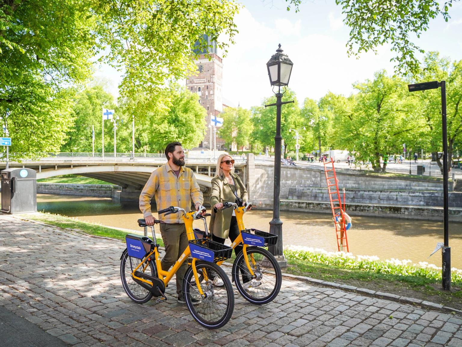 En mand og en kvinde står med gule lejecykler på en solrig sti ved floden med en bro og træer.