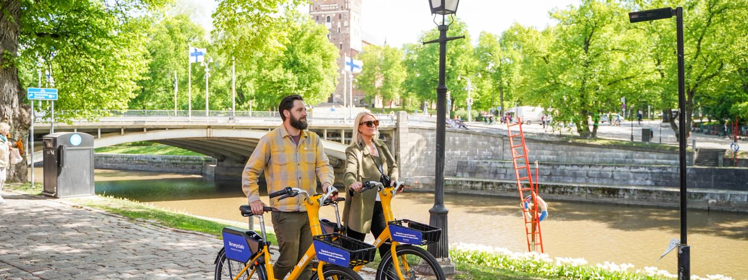 En mand og en kvinde står med gule lejecykler på en solrig sti ved floden med en bro og træer.