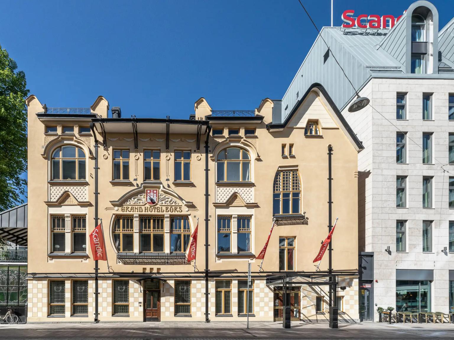 En udsmykket lysegul Grand Hotel Bours-bygning ved siden af et moderne Scandic-hotel under en blå himmel.