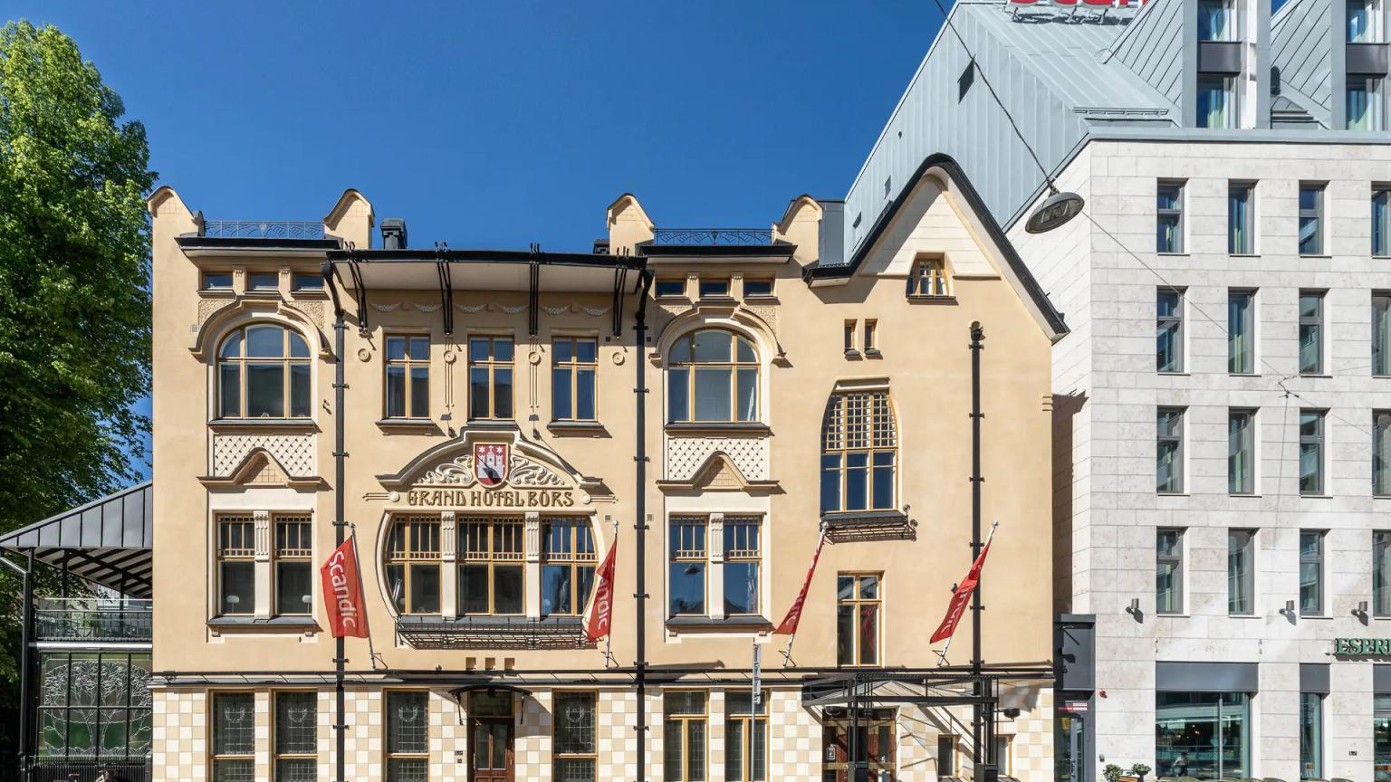 En udsmykket lysegul Grand Hotel Bours-bygning ved siden af et moderne Scandic-hotel under en blå himmel.