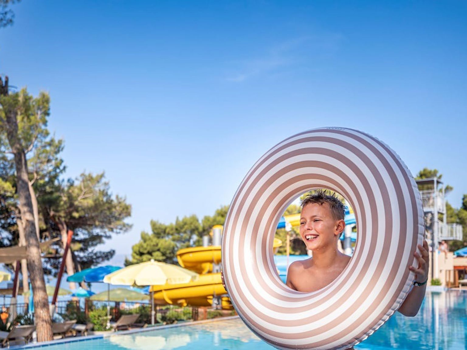En smilende dreng holder en stribet badering ved en pool med vandrutsjebaner på en solrig dag. Sunny Makarska by Valamar