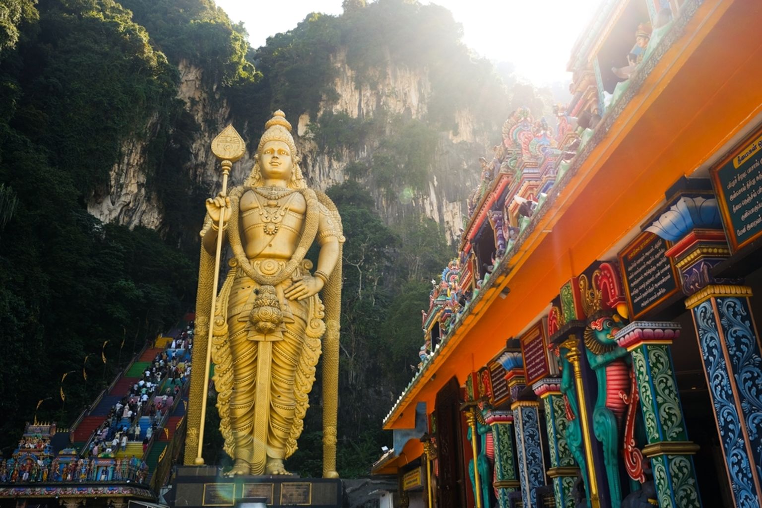 En massiv gylden statue af Lord Murugan står foran farverige trapper, der fører til Batu Caves, med en udsmykket tempelindgang til højre. Kuala Lumpur