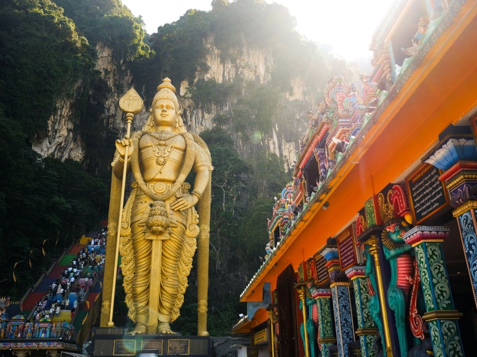 En massiv gylden statue af Lord Murugan står foran farverige trapper, der fører til Batu Caves, med en udsmykket tempelindgang til højre. Kuala Lumpur