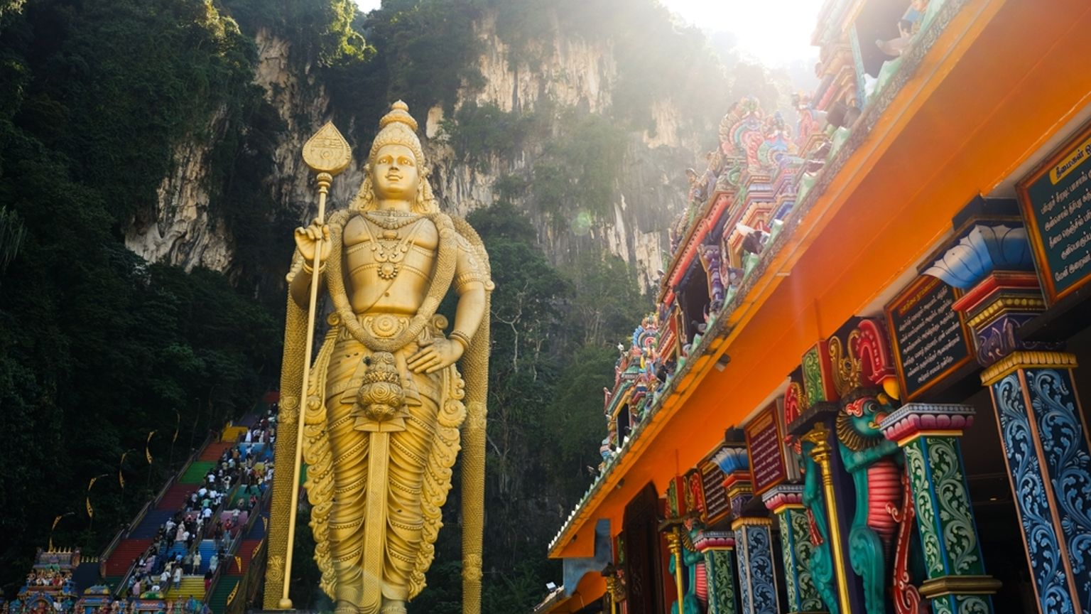 En massiv gylden statue af Lord Murugan står foran farverige trapper, der fører til Batu Caves, med en udsmykket tempelindgang til højre. Kuala Lumpur