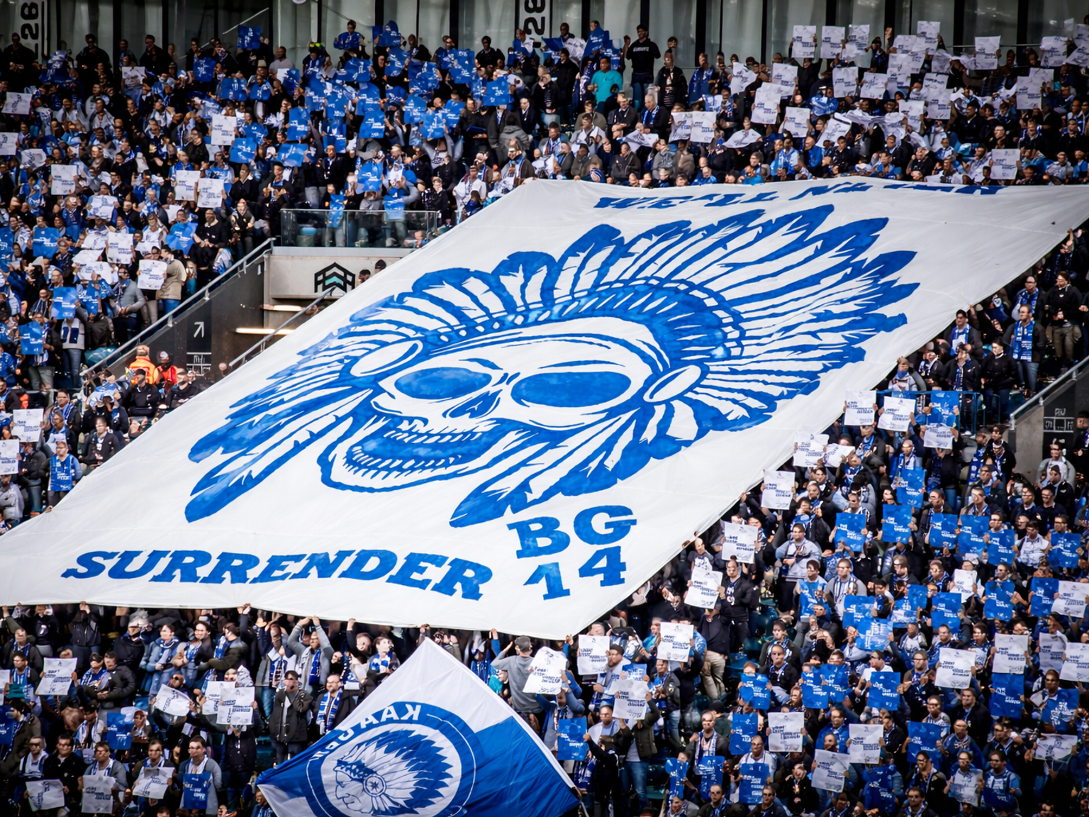 Stadionmenge, die ein großes weißes Banner mit einem blauen Totenkopf in einem gefiederten Kopfschmuck und dem Text „SURRENDER BG 14“ hält.