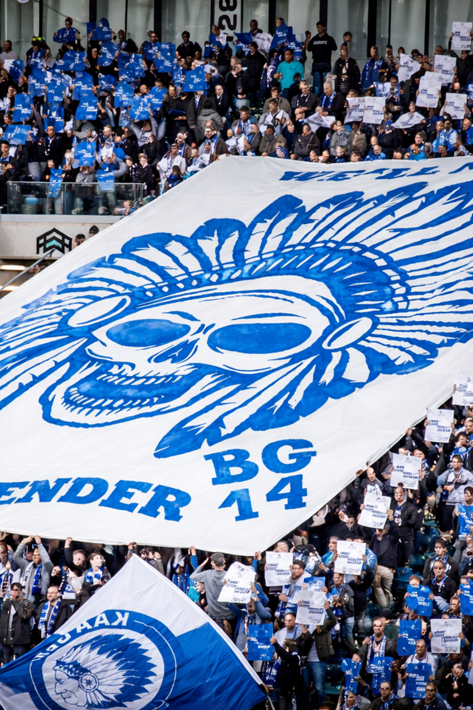 Stadionmenge, die ein großes weißes Banner mit einem blauen Totenkopf in einem gefiederten Kopfschmuck und dem Text „SURRENDER BG 14“ hält.