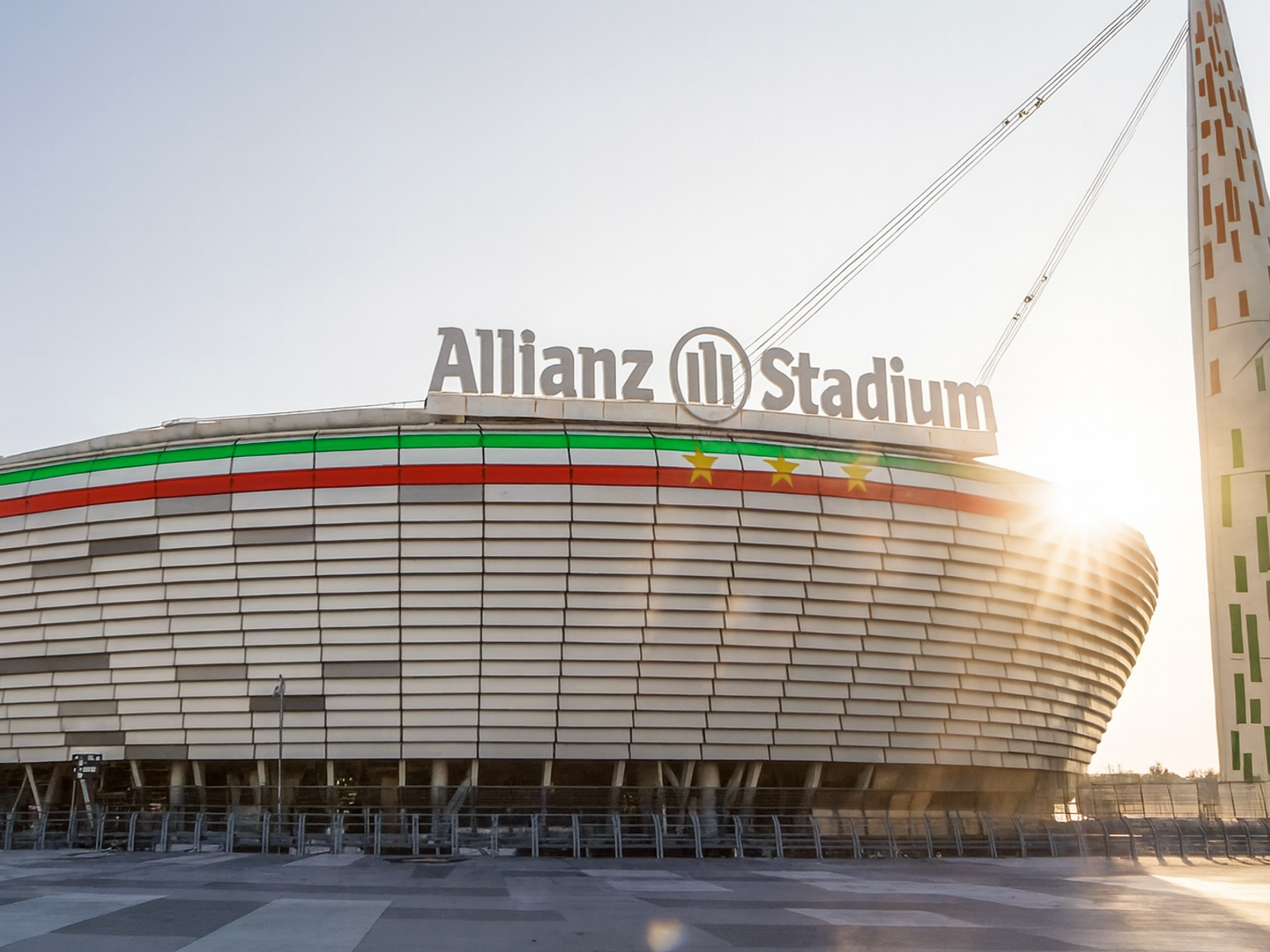 Modernes Allianz-Stadion-Äußeres mit rotem, weißem und grünem Streifen, goldenen Sternen und hellem Sonnenlicht.