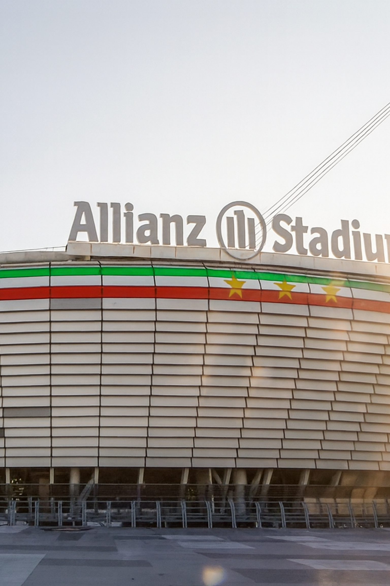 Modernes Allianz-Stadion-Äußeres mit rotem, weißem und grünem Streifen, goldenen Sternen und hellem Sonnenlicht.
