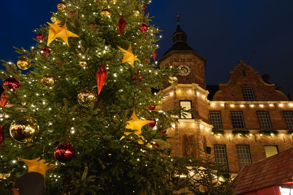 Sterkt opplyst juletre med røde og gullfargede ornamenter foran en murbygning med et klokketårn om natten.