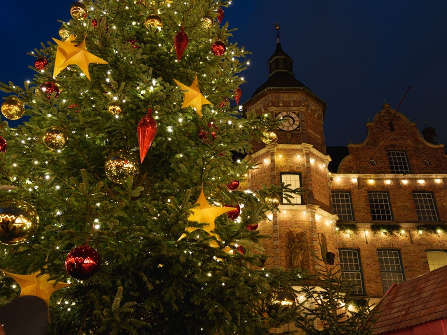 Sterkt opplyst juletre med røde og gullfargede ornamenter foran en murbygning med et klokketårn om natten.
