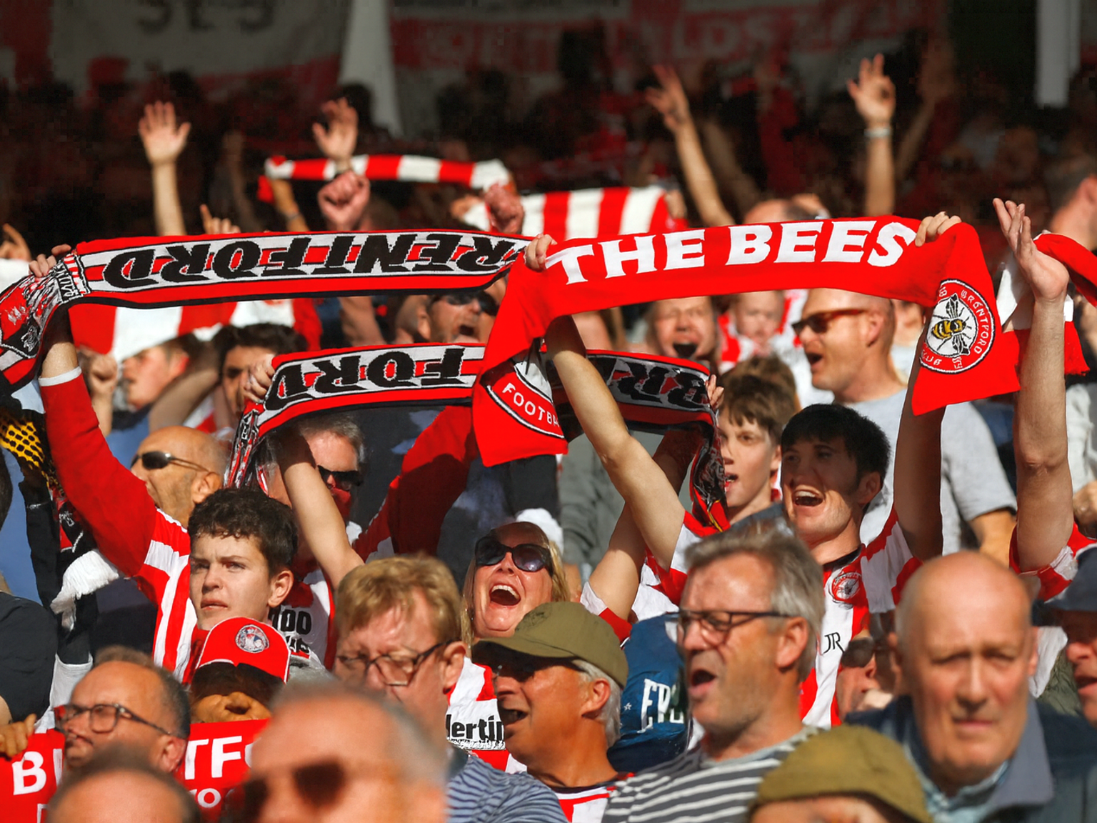 Les supporters de Brentford acclament, tenant des écharpes rouges et blanches avec les inscriptions "Brentford" et "The Bees."