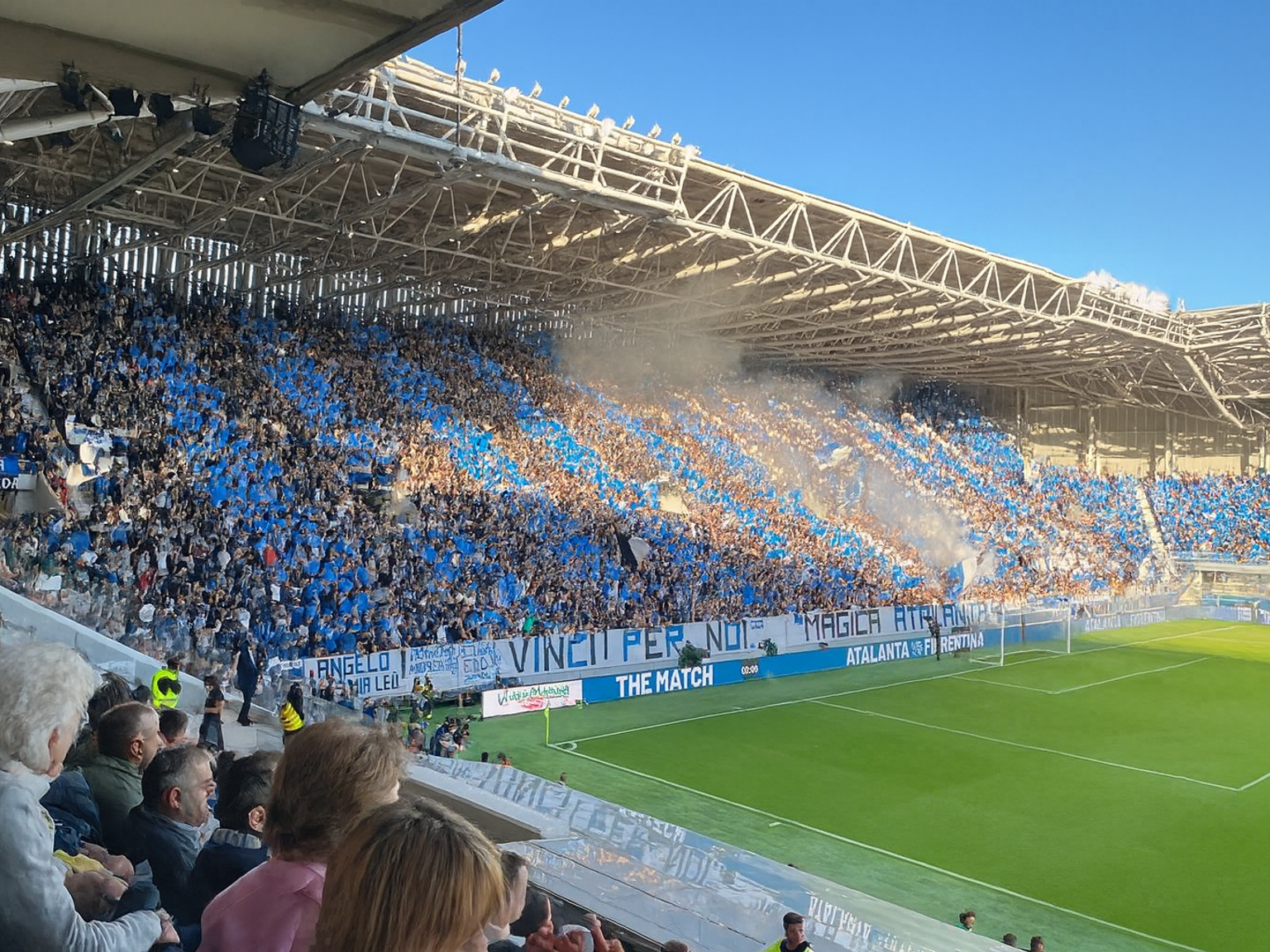 Täynnä oleva jalkapallostadion, joka on täynnä sinisiin pukeutuneita faneja, savua nousee katsomoista ja vihreä kenttä.