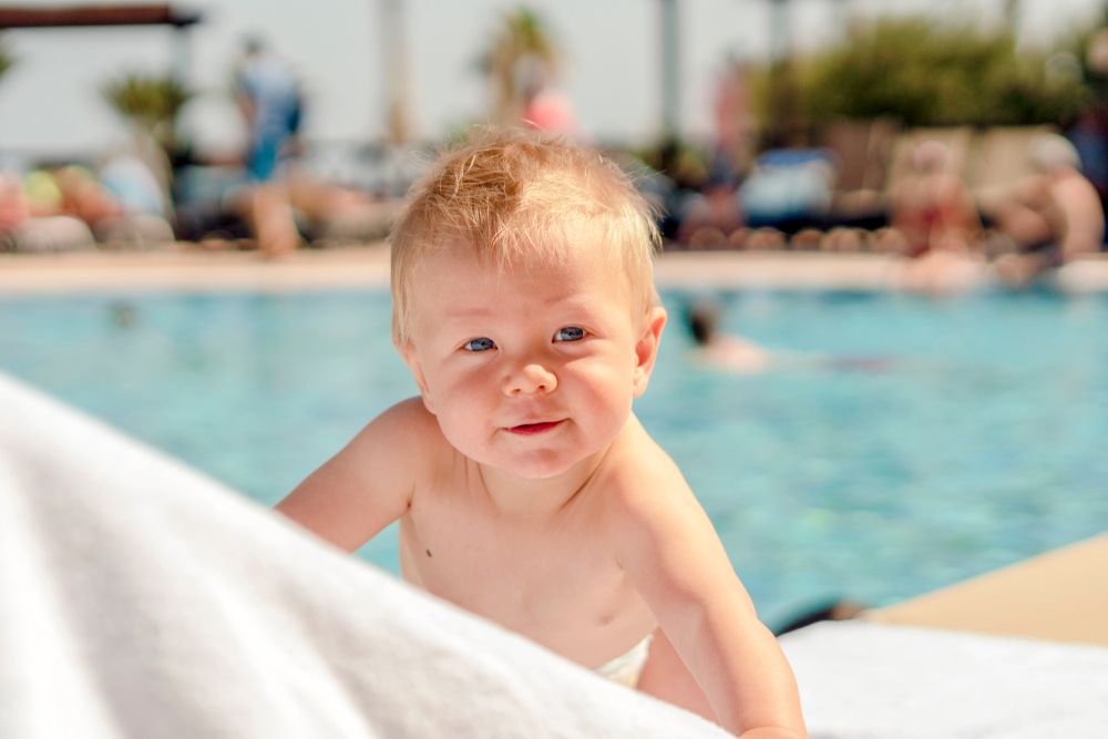 En smilende blond baby som titter over et hvitt håndkle ved et svømmebasseng.
