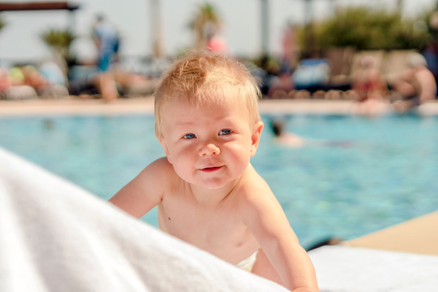 En leende blond bebis som kikar över en vit handduk vid en pool.
