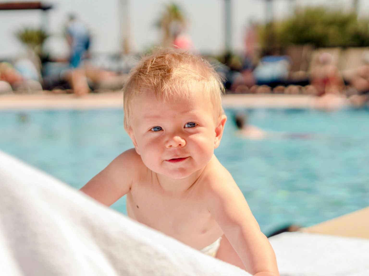 En smilende blond baby som titter over et hvitt håndkle ved et svømmebasseng.