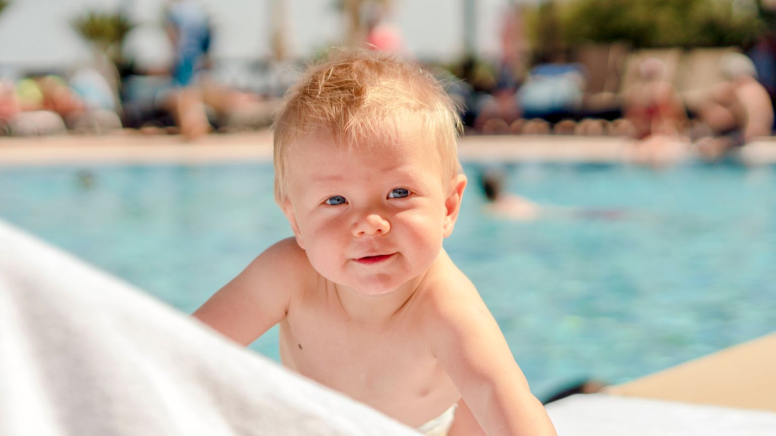 En leende blond bebis som kikar över en vit handduk vid en pool.