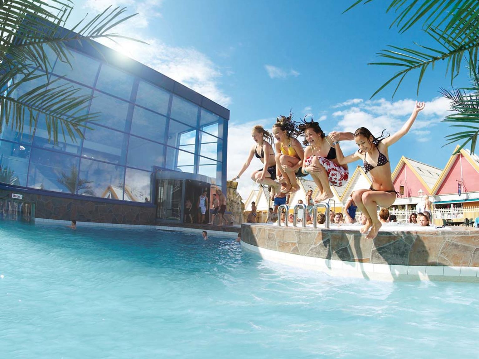 Fem unge kvinner hopper ned i et utendørs svømmebasseng på et feriested, med en stor glassbygning og fargerike hus i bakgrunnen. Lalandia Rødby