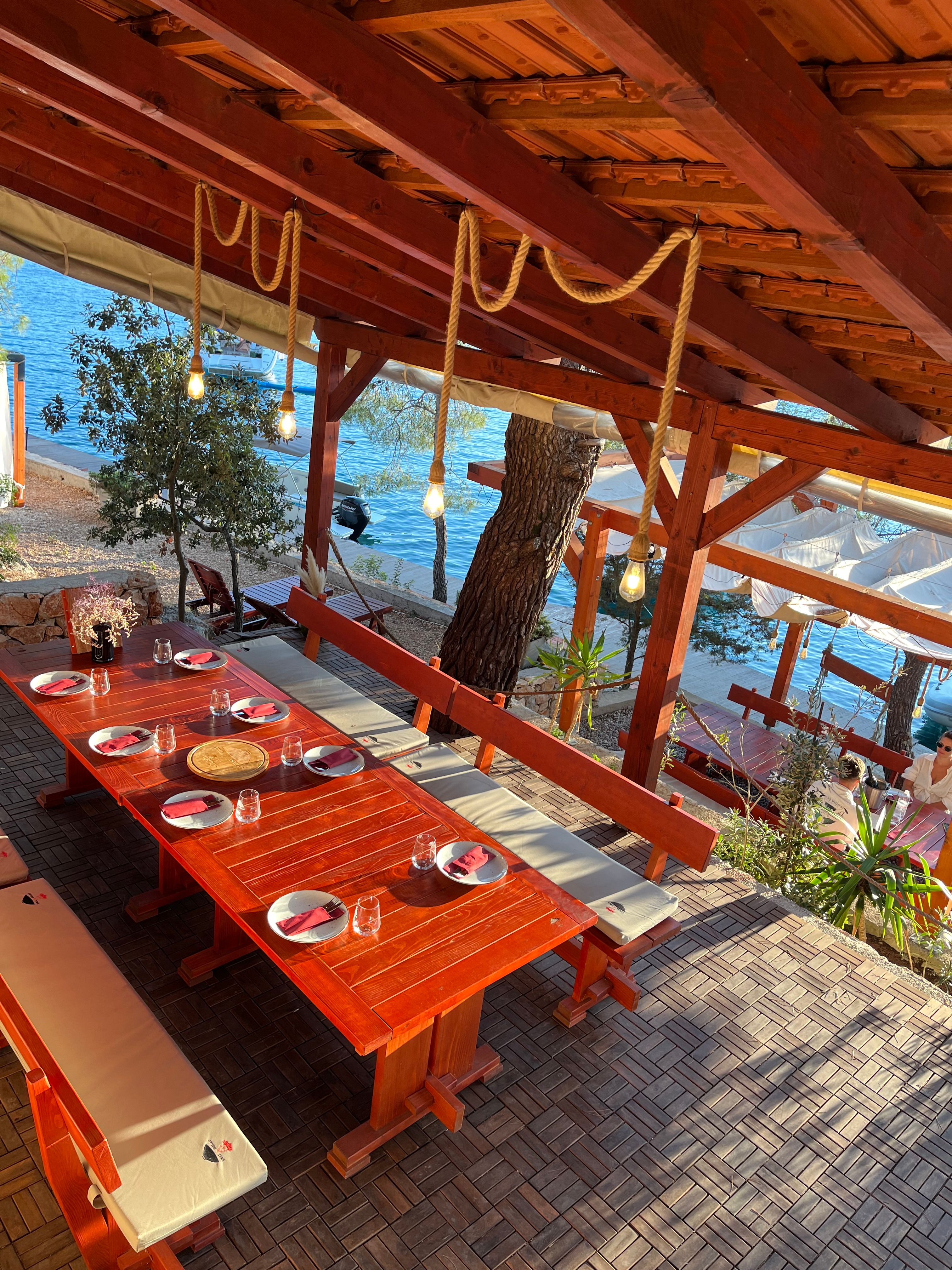 Espace de restauration en plein air avec une table rustique en bois dressée pour de nombreuses personnes, donnant sur la mer bleu clair en Croatie