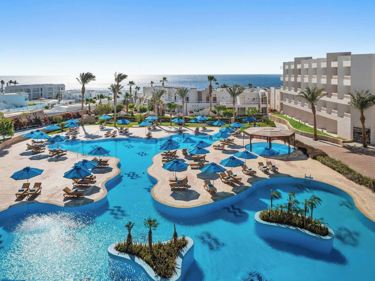 En stor, blå swimmingpool med solstolar och parasoller omgiven av palmer och vita resortbyggnader, med havet i bakgrunden.