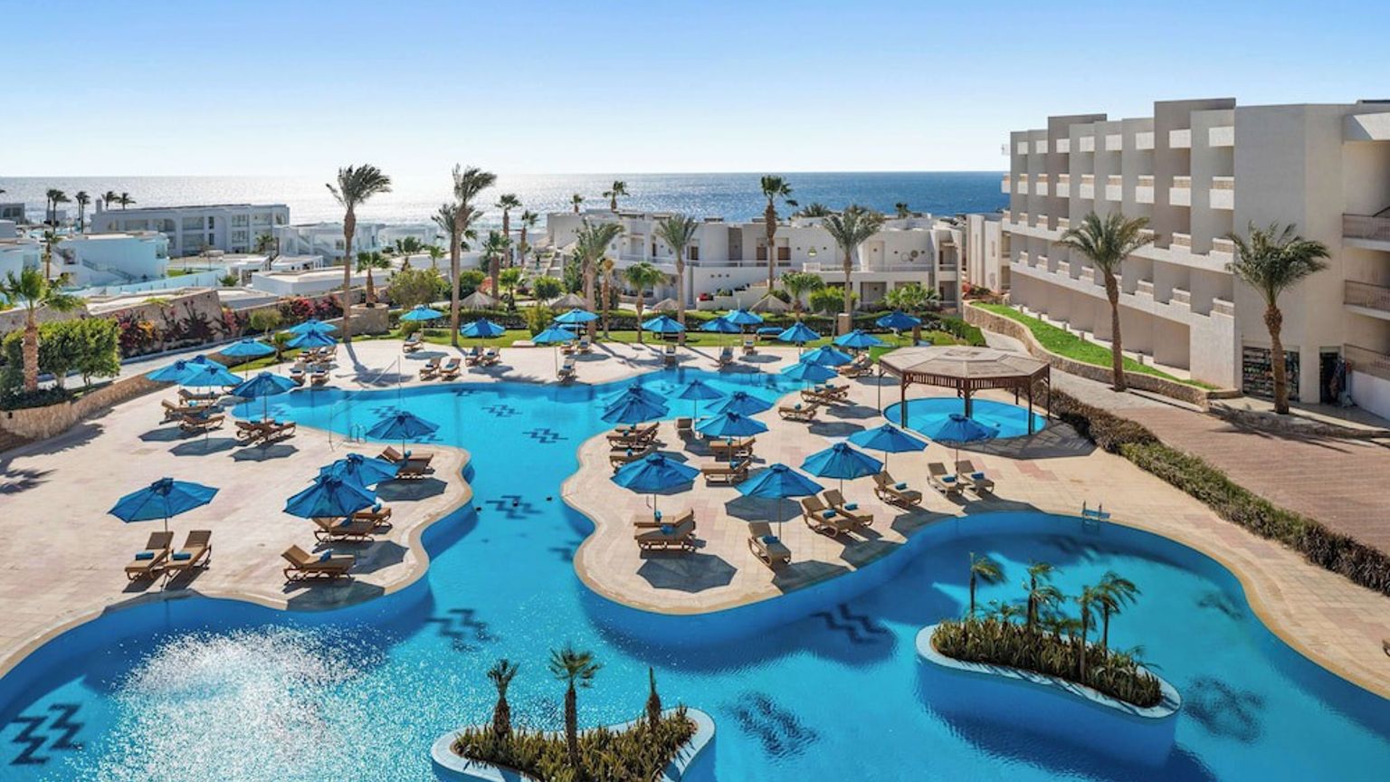 En stor, blå swimmingpool med solstolar och parasoller omgiven av palmer och vita resortbyggnader, med havet i bakgrunden.