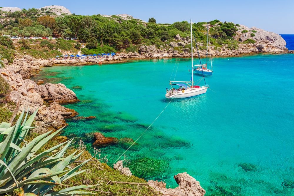 Two sailboats anchored in a clear turquoise bay with a rocky, green coastline. Rhodes, Greece