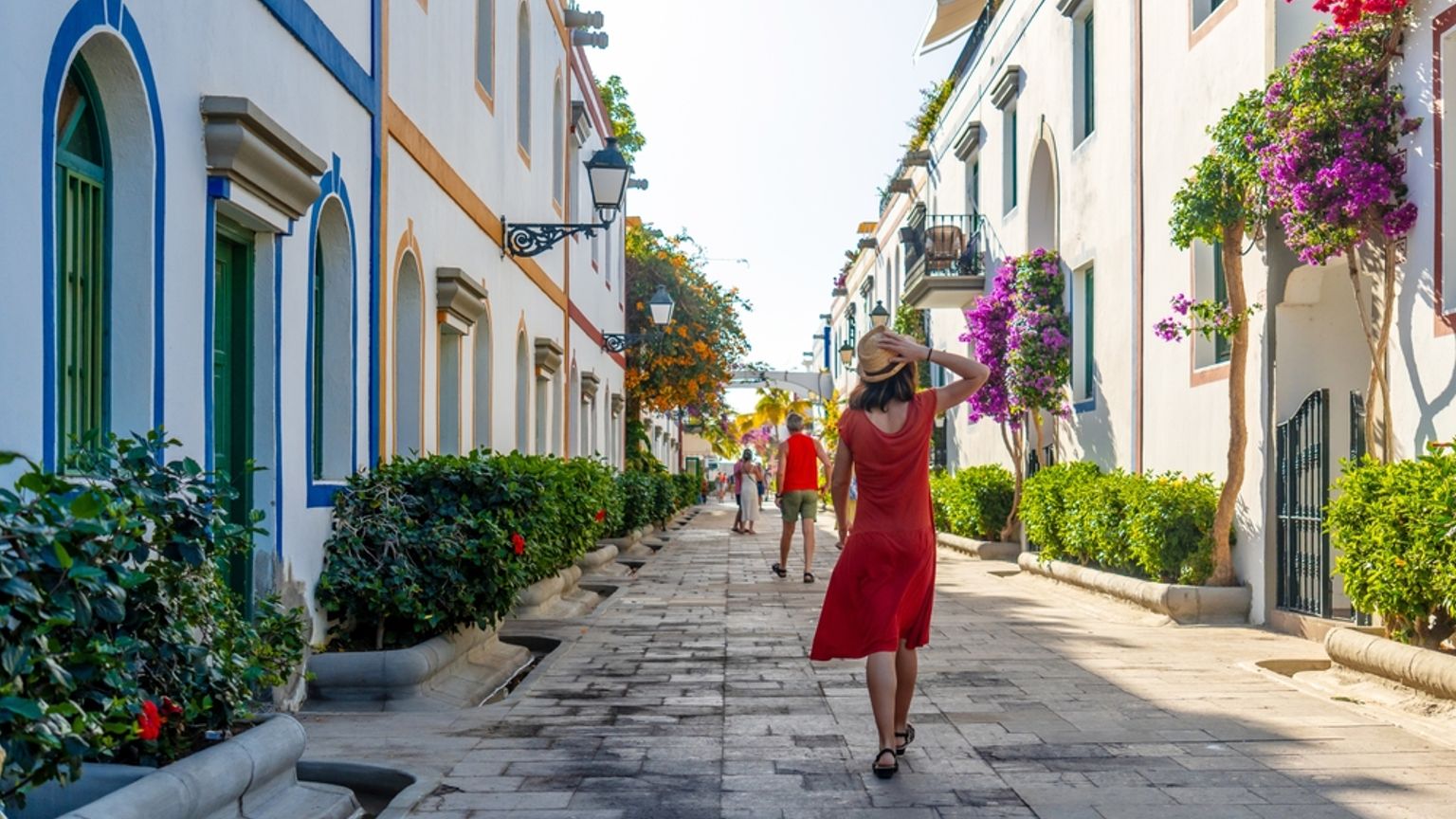 En kvinna i röd klänning och hatt går nerför en solig kullerstensgata kantad av vita byggnader och livlig bougainvillea.