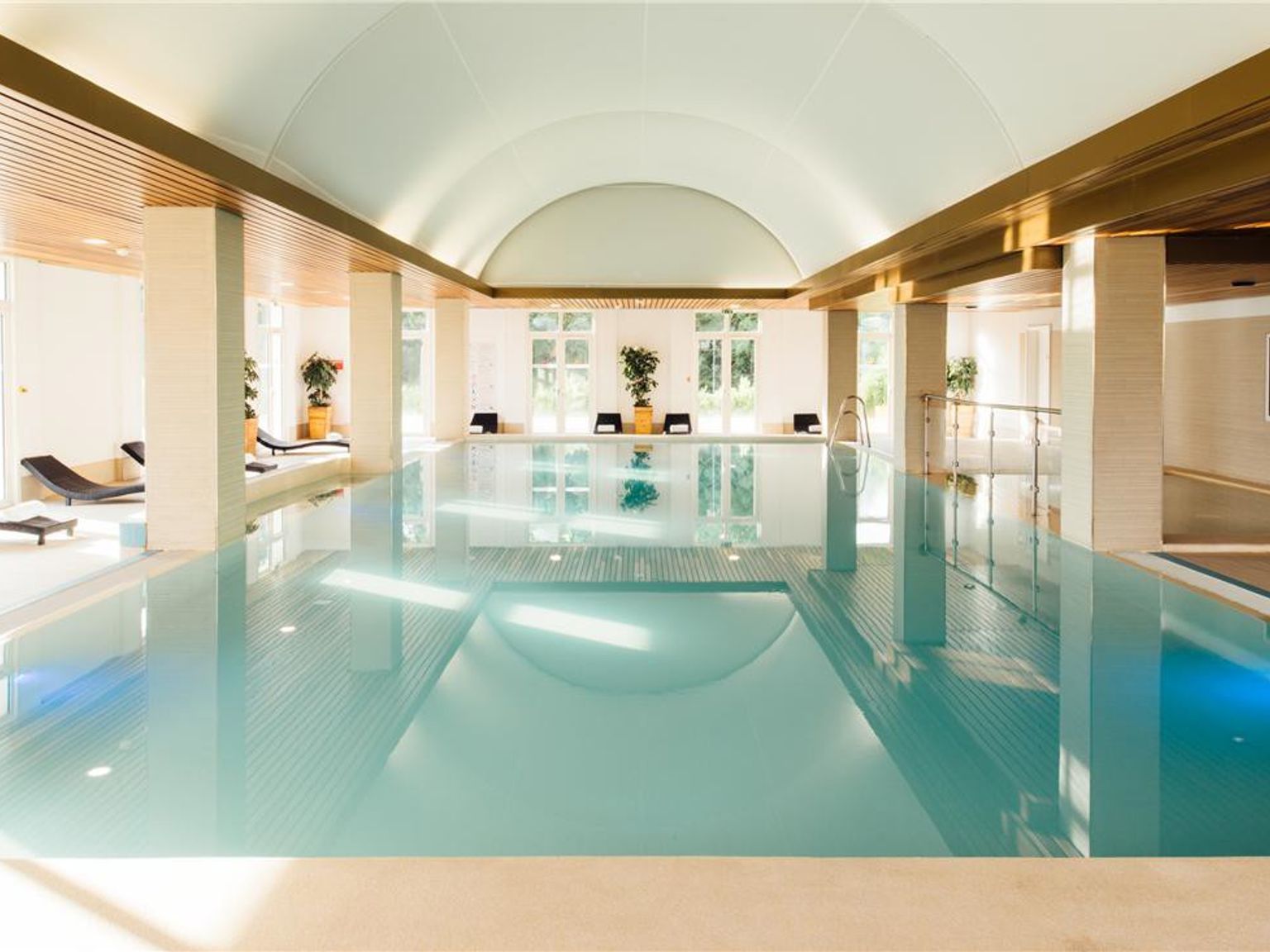 Et langt innendørs svømmebasseng med lyseblått vann, et hvelvet tak, store vinduer og solsenger langs sidene. Grand Magic Hotel