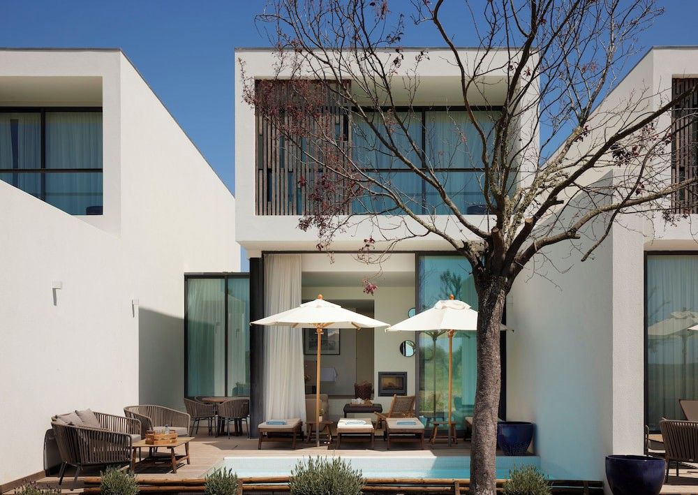 Modern vit arkitektur omsluter ett utomhusutrymme med en liten pool, solstolar och ett träd med ljusrosa blommor.