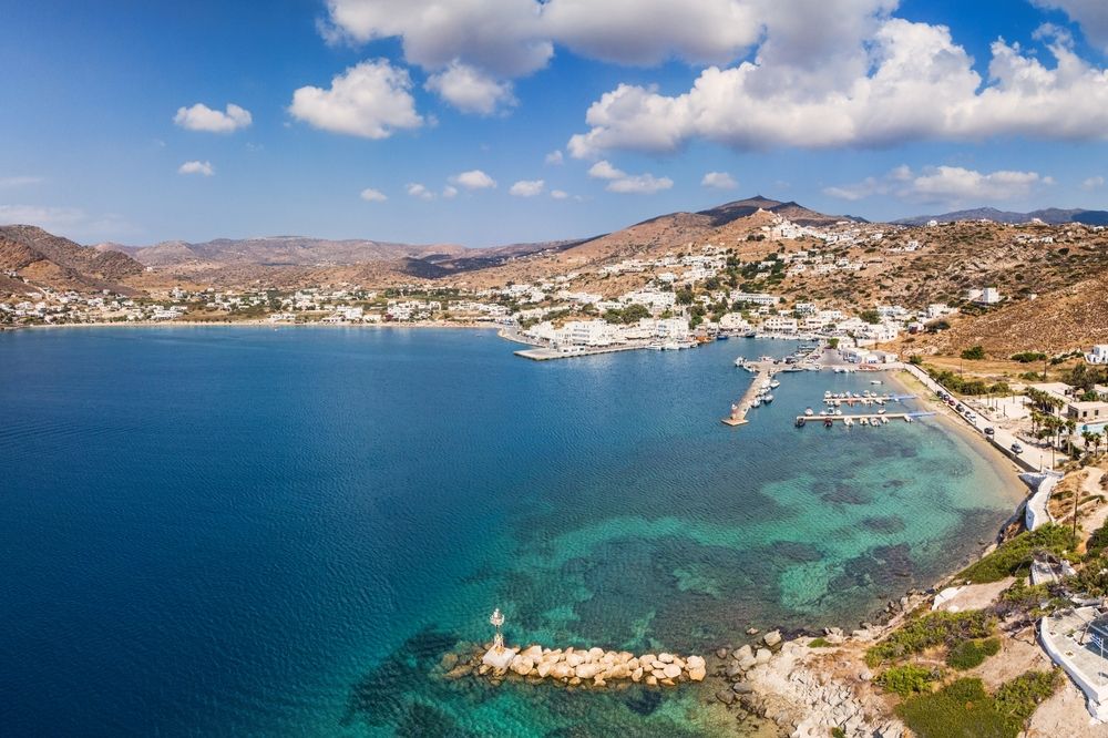 Aerial view of a coastal town with white buildings nestled in hills around a bay with a marina and clear blue water. Ios, Greece