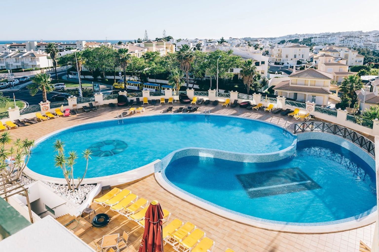 En flygbild av ett resortkomplex med två blå, sammanlänkade pooler omgivna av många gula solstolar och bostadsbyggnader i bakgrunden.